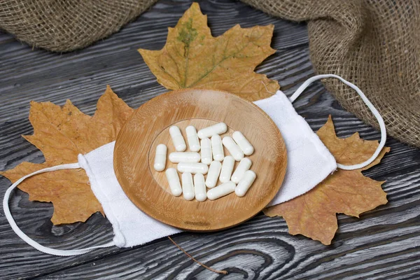 A handful of white pills on a wooden saucer. On a medical mask. Among the dried up maple leaves. On a pine board surface.