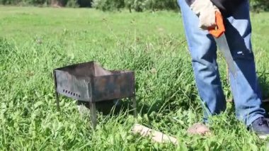 Maskeli bir adam barbekü için testereyi kesip odun kırıyor. Yakınlarda metal bir barbekü kutusu var. Salgın sırasında çayırda piknik.