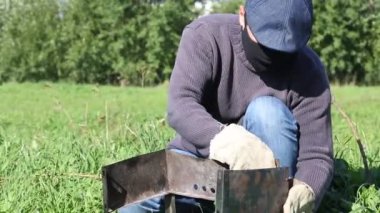 Maske ve eldiven takan bir adam metal bir ızgara kuruyor. Yan duvarları asıyor. Yakın planda çekilmiş. Salgın sırasında çayırda piknik..