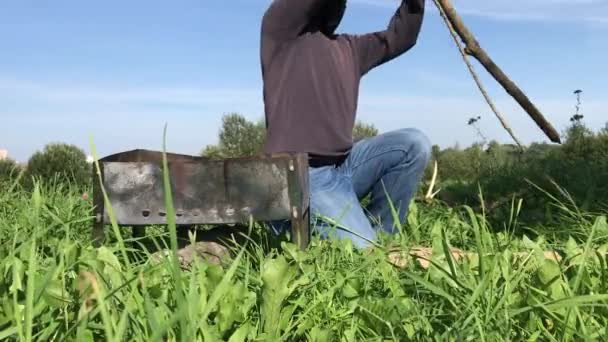 Muž v masce a rukavicích piluje a láme dřevo na gril. Nedaleko je umístěn kovový gril. Louka piknik během pandemie. — Stock video