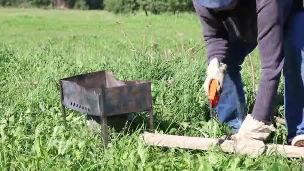 Un homme dans un masque et mitaines scies et casse le bois de chauffage pour le gril. Une boîte à barbecue en métal est installée à proximité. Un pique-nique dans la prairie pendant une pandémie. — Video