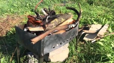 Yeşil bir çayırda metal brazier. Alevler ve duman görünür. Yakacak odun yakındaki çimlerin üzerinde yatıyor. Salgın sırasında çayırda piknik. Yakın çekim..