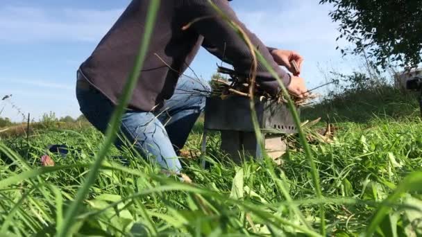 Muž zapálí suchou trávu a keře v kovovém grilu. Dřevo leží na trávě poblíž. Piknik na louce během pandemie. — Stock video