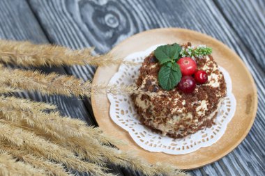 Tereyağlı kremalı pandispanya. Kızılcık ve nane ile süslenmiş. Yakınlarda kulakları olan kuru otlar var. Fırçalanmış çam ağaçlarının arka planında.