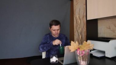 Adam video bağlantısı ile iletişim kuruyor. Tatlıyla kahve içer. Dizüstü bilgisayarın önündeki bir masada oturuyor. Arkasında gri bir arka plan örtüsü var..