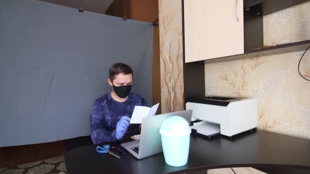 Un uomo in maschera e guanti comunica attraverso la comunicazione video mentre è seduto a un tavolo davanti a un computer portatile. Legge una lettera di carta e la mette in una busta. Stretched dietro di lui è un backgroun grigio — Video Stock