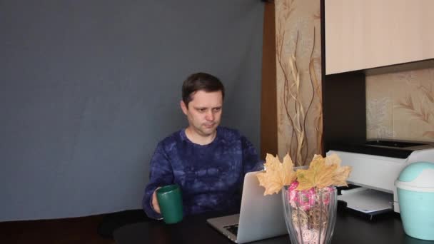 De man is aan het chatten via een videolink. Hij neemt koffie en dessert en eet. Zit aan een tafel voor een laptop. Achter hem ligt een grijze achtergrond doek.. — Stockvideo