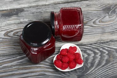 Cam kavanozlarda ahududu reçeli. Yakınlarda bir fincan tabağında ahududu var. Ahşap tahtaların üzerinde güzel bir dokusu var..