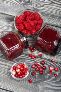 Cam kavanozlarda kızılcık reçeli. Konteynırlarda yabanmersini ve ahududu var. Bazı meyveler yüzeye saçılmıştır. Ahşap tahtaların üzerinde güzel bir dokusu var..