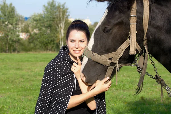 The girl strokes and feeds the horse. The horse grazes on a green meadow.