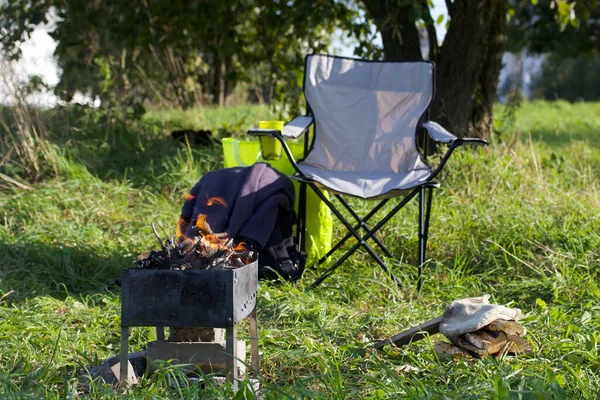 Camping chair and metal grill with a light fire. They are standing in a green meadow. Autumn picnic.