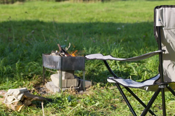 Camping chair and metal grill with a light fire. They are standing in a green meadow. Autumn picnic.