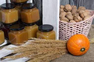 Kavanozda meyve reçeli. Beyaza boyanmış ahşap bir kutuda duruyor. Yanında bir sepet ceviz, bir portakal kabağı ve bir buket kuru ot var..