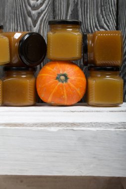 Kavanozda meyve reçeli. Yakınlarda turuncu bir balkabağı var. Beyaz boyalı ahşap bir kutunun üzerinde duruyor. Siyah çam ağaçlarının arka planında.