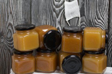 Kavanozda meyve reçeli. Tıbbi bir maske takıyor. Beyaz boyalı ahşap bir kutunun üzerinde duruyor. Siyah çam ağaçlarının arka planında.