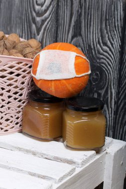 Bir sepet ceviz ve reçel. Yakınlarda turuncu bir balkabağı var. Tıbbi maske takıyorlar. Beyaz tahta bir kutunun üzerinde duruyorlar..