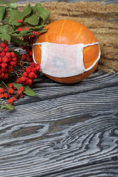 Orange pumpkin in a medical mask. Lies on black pine boards. Nearby is a bouquet of dried grass and mountain ash.