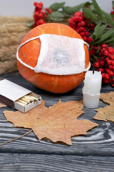 Orange pumpkin in a medical mask. Lies on black pine boards. Nearby is a bouquet of dried grass and mountain ash. There is a candle stub.