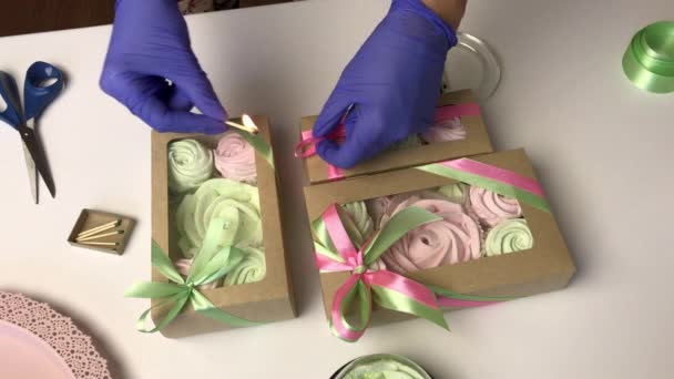 Жінка упаковує рожевий і світло-зелений зефір в подарункову упаковку. Зв'язує їх стрічкою і спалює їх сірниками . — стокове відео