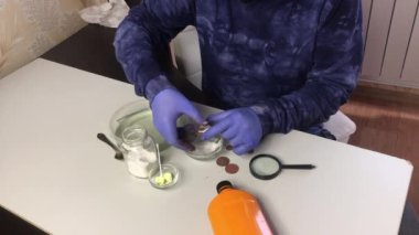 Maskeli ve lastik eldivenli bir adam bakır paraları soda ve suyla siler. Yakınlarda bakır sikkeleri temizlemek için fosforik asit ve sülfürik merhem var.