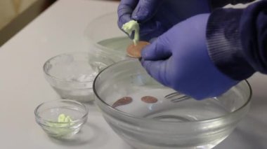 Lastik eldivenli bir adam sikkeye sülfür merhemi sürer. Bir madeni parayı sıcak suya sürter. Sülfürik merhemle bakır paraların şekli.