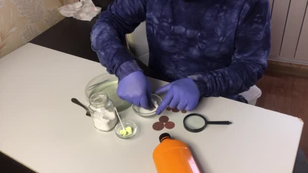 Un hombre con una máscara y guantes de goma limpia monedas de cobre con una papilla de refresco y agua. Cerca hay ácido fosfórico y ungüento sulfúrico para limpiar monedas de cobre. — Vídeos de Stock