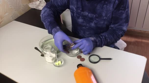 Een man met een masker en rubberen handschoenen veegt koperen munten af met een korrel frisdrank en water. In de buurt is fosforzuur en zwavel zalf voor het reinigen van koperen munten — Stockvideo