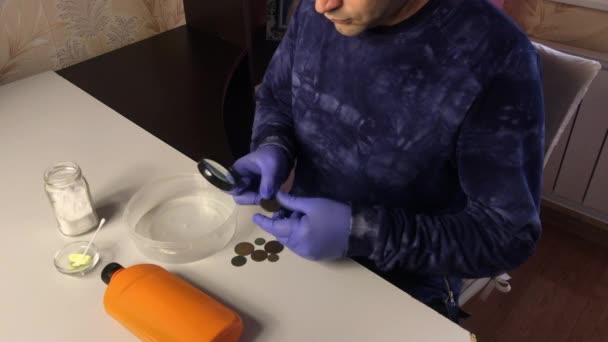 A man in rubber gloves examines coins through a magnifying glass. Chemicals for cleaning copper coins are scattered nearby. — Stock Video