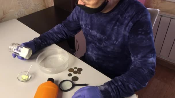 A man wearing a mask and rubber gloves examines chemicals for cleaning copper coins. Coins covered with corrosion are being cut on the table nearby. — Stock Video