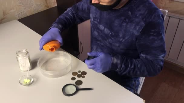 A man wearing a mask and rubber gloves pours orthophosphoric acid into a container. Corroded coins and chemicals for cleaning copper coins are being cut on the table nearby. — Stock Video