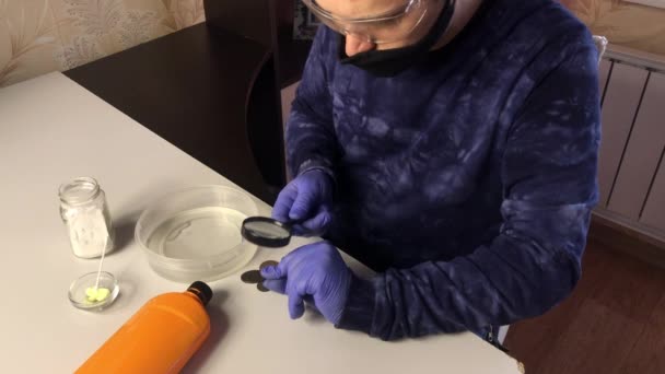 Un homme portant un masque et des gants de caoutchouc met des pièces de monnaie dans l'acide phosphorique. Des pièces de monnaie et des produits chimiques corrodés pour nettoyer les pièces de cuivre sont coupés sur la table à proximité. — Video