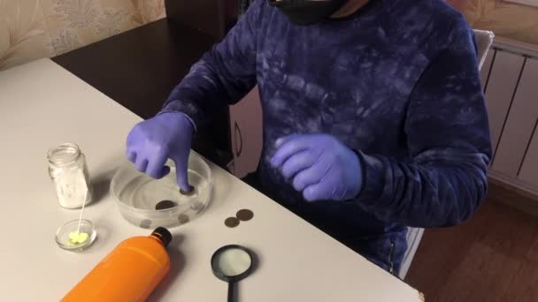 Un homme portant un masque et des gants de caoutchouc met des pièces de monnaie dans l'acide phosphorique. Des pièces de monnaie et des produits chimiques corrodés pour nettoyer les pièces de cuivre sont coupés sur la table à proximité — Video