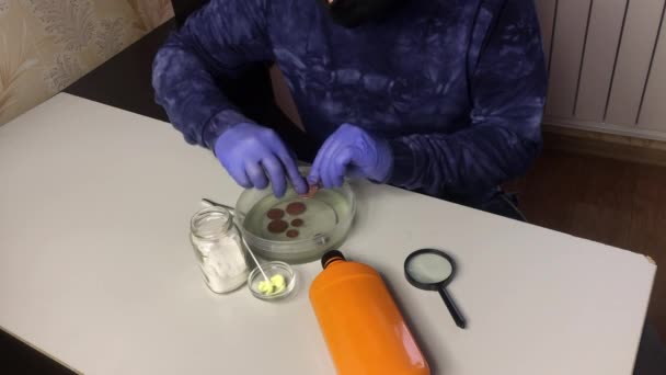 Ein maskierter Mann mit Gummihandschuhen steckt Münzen in Phosphorsäure und kontrolliert den Reinigungsprozess. Plaque mit den Fingern reiben. — Stockvideo