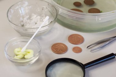 Masanın yüzeyinde soyulmuş bakır paralar var. Aşınmış madeni paralar fosforik asitle dolu bir kapta yakınlarda duruyor. Patinaj için sülfürik merhem.