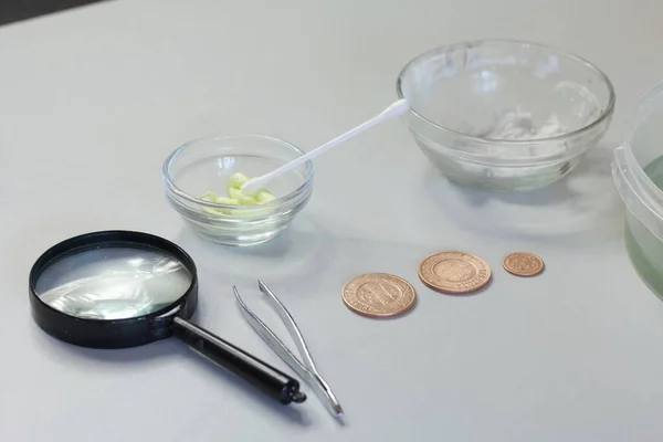 Peeled copper coins lie on the table surface. Corroded coins are lying nearby in a container with phosphoric acid. Sulfuric ointment for patination.