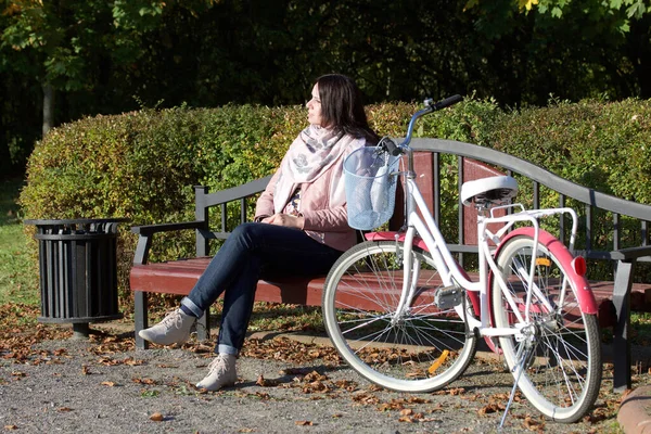 Sonbahar parkında bisiklet süren bir kız. Bankta oturuyor. Yakınlarda bisiklet var..