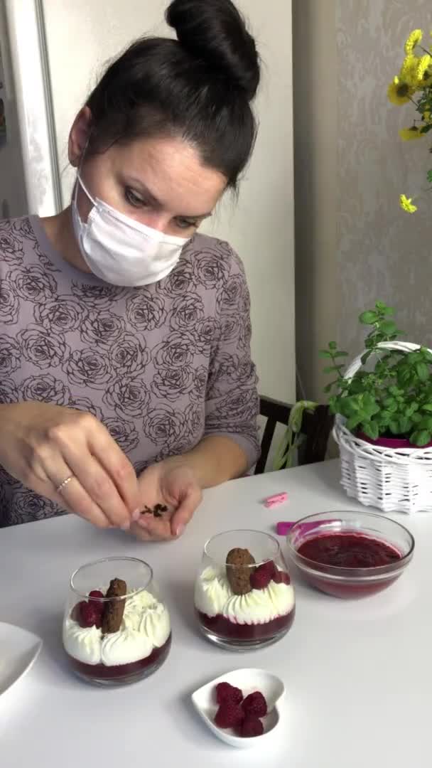 Женщина в медицинской маске украшает десерт в стакане шоколадной крошкой. Десерт с малиновым желе. Ингредиенты выложены на поверхность стола. Украшенный кухней — стоковое видео