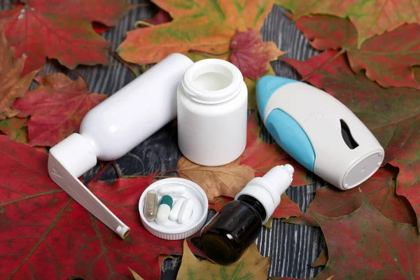 Pill container, inhaler and nasal drops. The lid of the container and several tablets are next to each other. Among the autumn maple leaves on the black pine boards.