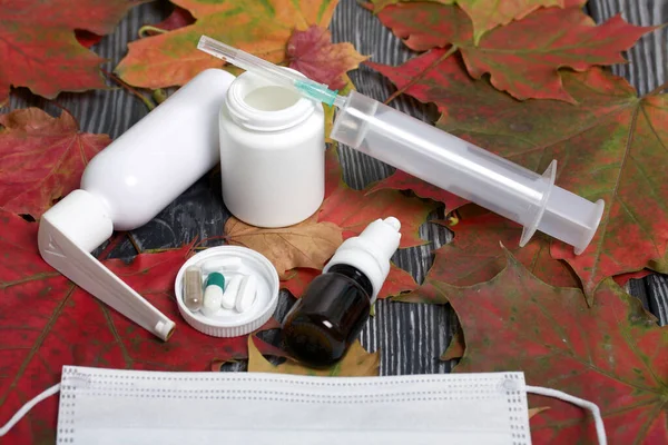 Medical mask, container with pills and other drugs. The lid of the container and several tablets are next to each other. Among the autumn maple leaves on the black pine boards.