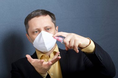 Takım elbiseli, kravatsız bir adam. Yüzünde tıbbi maske var. Bir sprey şişesinin antiseptikle nasıl çalıştığını gösteriyor. Önünde aydınlatma için lambası olan bir akıllı telefon standı var. Video iletişimi.