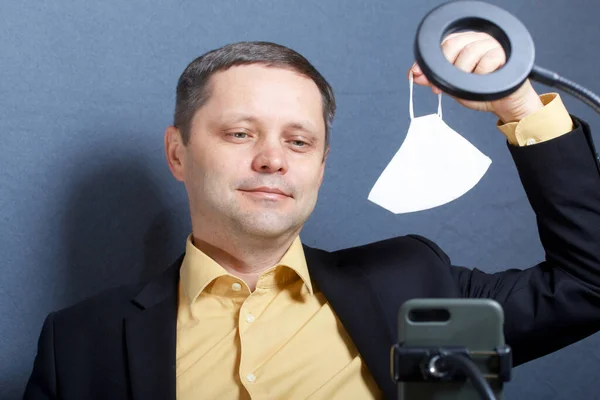 A man in a suit without a tie. Demonstrates a medical mask on camera. In front of him is a stand for a smartphone with a lamp for illumination. Video communication.