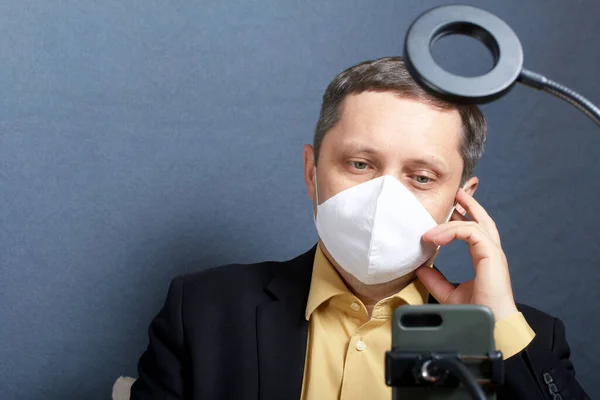 ネクタイのないスーツの男 顔に医療マスク 彼の前には照明用のランプを備えたスマートフォンのスタンドがあります ビデオ通信 — ストック写真