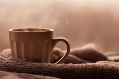 Buharlı kahve fincanı ve bulanık arka planda örülmüş süveter. Bej renkler, sonbahar kupası, sonbahar kahvesi. Metin eklemek için afiş alanı modeli.