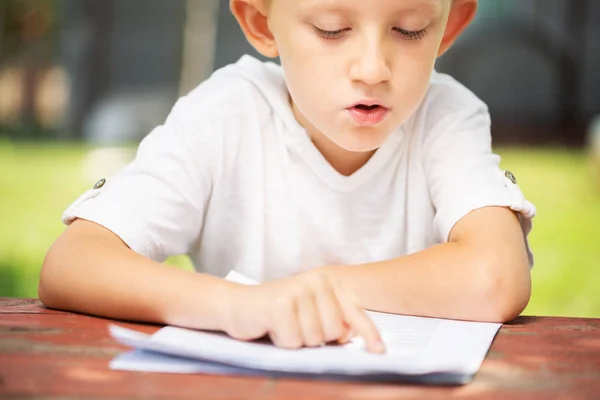 白いTシャツを着た小さな男の子が学校の概念に戻って屋外で読書しています. — ストック写真