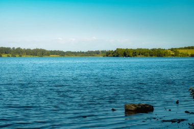 Göl kenarından çevre orman manzarasına güzel bir manzara, arka planda ormanın manzarası ve ön planda bir kaya daha fazla derinlik katıp fotoğrafa alıkoymak. Sabah ilk iş doğa yürüyüşüne çıkmak için güzel bir yol.
