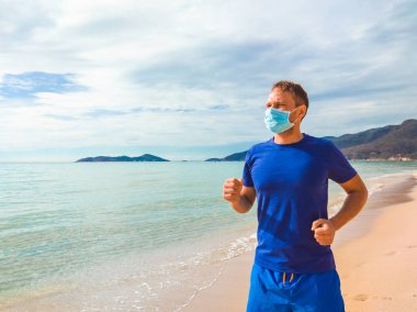 Coronavirus COVID 19. Genç ve güzel bir adam sabah karantina sırasında deniz kenarında spor egzersizleri yapıyor ve koşuyor. Yüz maskesi korumalı cerrahi sterilize yaşam