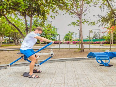 Fiziksel egzersiz yapan, şınav çeken, güç egzersizi yapan boş şehir parkında metal bir spor salonuna yaslanan ve Covid19 önleme için maske takan bir adam. Karantina ve koronavirüste sağlıklı yaşam tarzı