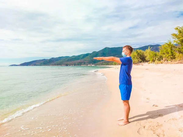 Coronavirus COVID 19. Joven hombre hermoso haciendo deporte realizar ejercicios de ejercicios y ponerse en cuclillas cerca del mar por la mañana durante la cuarentena. Vida activa en la protección de mascarillas quirúrgicas esterilizantes — Foto de Stock