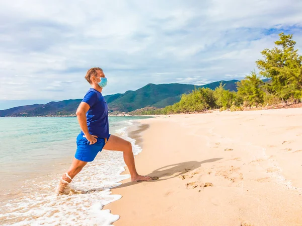 Coronavirus COVID 19. Pemuda tampan yang melakukan olahraga melakukan latihan dan berjongkok di dekat laut di pagi hari selama karantina. Aktif hidup dalam bedah sterilisasi wajah topeng perlindungan — Stok Foto