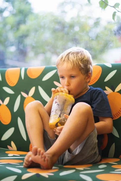 Front visning af glad tørstig blond dreng drikker limonade, frugt te, kombucha is te med fersken og citron, wellness appelsin drikkevarer fra plastik kop med strå, sidder på multi farve sofa. Lodret - Stock-foto